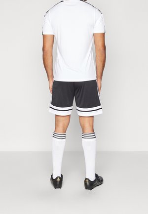 Ein Mann steht mit dem Rücken zur Kamera, trägt ein weißes Sport-T-Shirt, schwarz-weiß gestreifte Shorts, weiße knielange Socken und schwarze Stollenschuhe.
