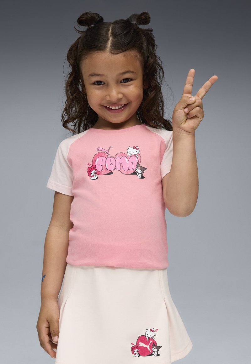 Uśmiechnięta dziewczynka z kręconymi włosami w różowej koszulce Puma Hello Kitty i jasnej spódnicy pokazuje znak pokoju na szarym tle.