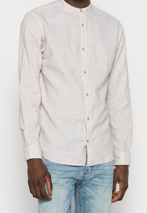 Homme portant une chemise beige clair à manches longues avec des boutons en bois et un jean bleu clair sur un fond uni.