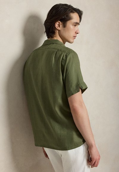 Jeune homme de profil portant une chemise à manches courtes vert olive et un pantalon blanc devant un mur beige uni.