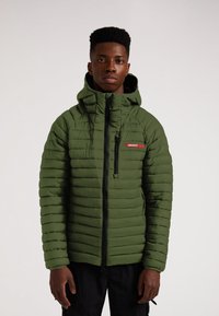 Grön puffjacka med horisontell quiltad design, framdragkedja och huva. Har röd "JACKIT" logotyp på bröstfickan.