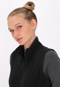 Vest din fleece negru cu guler înalt și închidere cu fermoar, purtat peste o bluză gri cu mânecă lungă; textură netedă și design ajustat.