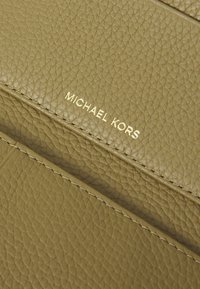 Portfel z fakturowanej skóry w oliwkowym kolorze, z złotym logo "MICHAEL KORS" i widocznym szwem. Kompaktowy design z wieloma slotami na karty.