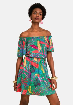 Femeie care poartă o rochie colorată, cu umerii goi, cu motive de frunze tropicale și păsări, accesorizată cu cercei cu flori roz și brățări aurii.