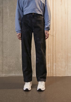 Person, die ein hellblaues Langarmhemd, schwarze Hosen mit weitem Bein und weiße Turnschuhe trägt, steht auf einem dunkel gesprenkelten Boden.