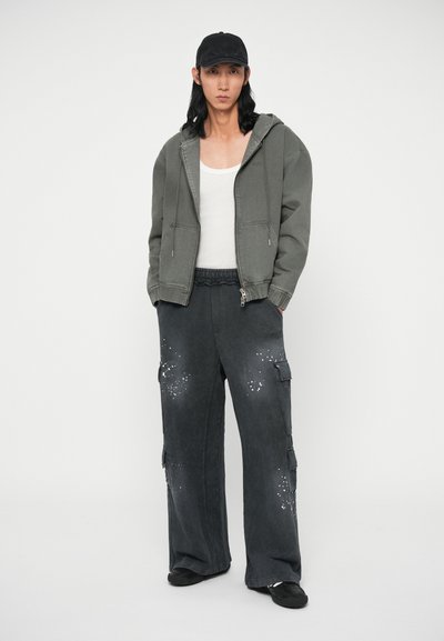 Šedá kapucňová bunda, biele tielko, tmavé cargo nohavice s dekoratívnym rozliatím, čiapka čiernej farby a čierne topánky. Hladká tkanina, voľný strih.