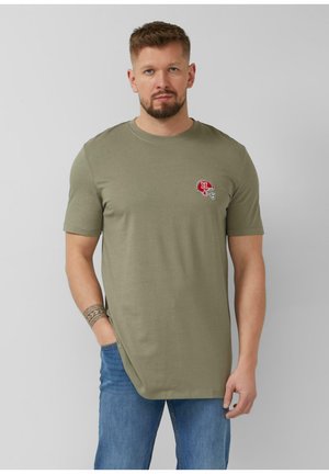 Homme barbu portant un T-shirt vert olive avec un logo de casque de football rouge, un jean bleu et un tatouage au poignet à motifs, debout devant un fond gris.