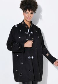 Chaqueta de ante negro con recortes circulares y acentos metálicos, mangas largas, cuello alto y un diseño de corte holgado.