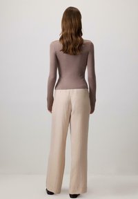 Woman with wavy brown hair, wearing a fitted long-sleeve taupe top and loose light beige pants, standing facing a plain white wall.