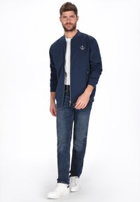 Marineblå bomberjakke lavet af glat stof, med lynlås foran og sidelommer, kombineret med lyse denimjeans og hvide sneakers.