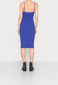 Robe midi côtelée bleu royal avec bretelles spaghetti, silhouette ajustée et fente sur le côté. Portée avec des bottines noires à talons épais.