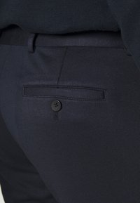 Tmavě modré kalhoty s hladkým povrchem, obohacené o zadní kapsu uzavřenou černým knoflíkem a jemným designem pasu.