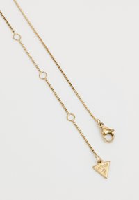 Gouden ketting met rechthoekige schakels, voorzien van ronde accenten en een driehoekig label met de tekst "GUESS." Klauwsluiting voor een veilige sluiting.