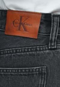 Blugi negri din denim, cu etichetă maro din piele imprimată cu „Calvin Klein” și „CK”. Detalii notabile de cusături și design standard al buzunarelor.