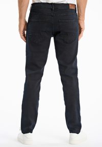 Jeans de color azul oscuro con un ajuste slim, fabricados en denim. Presentan dos bolsillos traseros y un parche de cuero marrón en la cinturilla. Zapatillas blancas visibles.