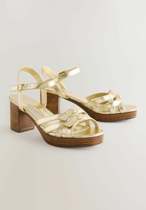 Auksinės metalinės basutės su mediniu blokiniu kulnu, su kryžminėmis dirželiais ir sagtelėmis. Lygi tekstūra su atvirų pirštų dizainu.