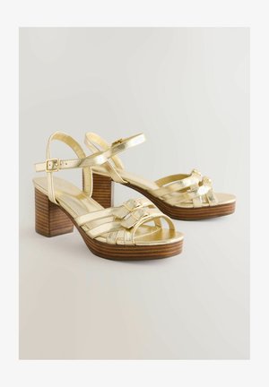 Goldfarbene, metallische Sandalen mit hohen Absätzen aus Holz, die über kreuzverlaufende Riemen und Schnallenakzente verfügen. Glatte Textur mit einem offenen Zehendesign.