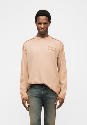 Jeune homme debout, portant une chemise beige à manches longues avec un texte orange et un jean bleu délavé, sur un fond blanc uni.
