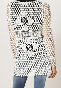 Vit virkad cardigan med blommönster, öppen stickning, långa ärmar och scalloped fåll. Matchas med blå jeans.