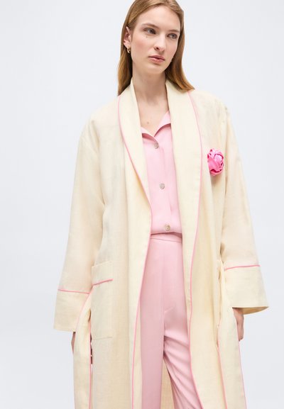 Γυναίκα που φοράει ρομπάκι εκρού με ροζ περιθώριο πάνω από ροζ πουκάμισο με κουμπιά και παντελόνι, με ένα ροζ υφασμάτινο λουλούδι στην τσέπη της ρόμπας.