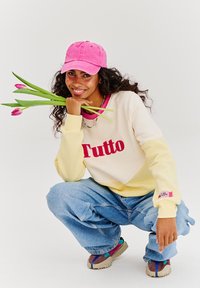 Bílá a žlutá mikina s nápisem "Tutto" v červené, světle modré volné džíny, růžová kšiltovka, barevné tenisky a tulipány v ruce.