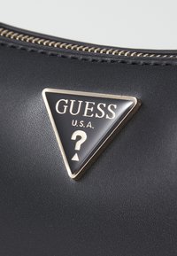 Black textured material with a triangular gold-accented GUESS logo featuring a question mark. Zipper detail along the top edge.