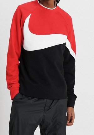 Person mit einem rot-weiß-schwarzen Nike-Sweatshirt mit großem Swoosh-Logo und schwarzen Nike-Trainingshosen vor neutralem Hintergrund.