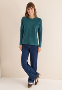 Turkusowy sweter z dzianiny, z okrągłym dekoltem, w zestawieniu z ciemnoniebieskimi spodniami o szerokich nogawkach i brązowymi sneakersami z białymi akcentami.