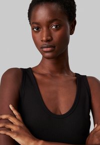 Canotta nera a coste con scollatura a V profonda e spalline larghe, che mette in risalto una texture liscia e un design aderente. Tono della pelle visibile sulle braccia.