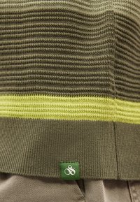 Κοντινό πλάνο πλεκτού υφάσματος σε ελιά πράσινο με ραβδωτή υφή, κίτρινη ρίγα και ένα μικρό πράσινο ταμπελάκι με λευκό λογότυπο στο τελείωμα.