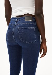 Jean stretch bleu foncé à taille haute, avec deux poches arrière et une étiquette noire gravée "ARMEDANGELS" sur la ceinture.