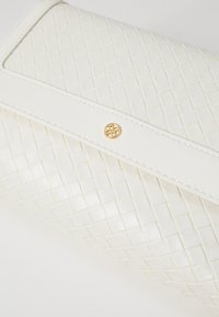 Hvit vevd skinnclutch med en glatt, strukturert klaff og en gullfarget emblemdetalj som fremhever en kviltet tekstur.