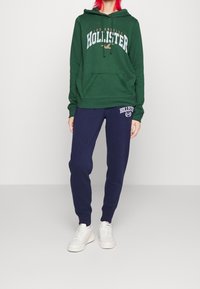Sudadera verde con capucha y logo de "Hollister", con bolsillo delantero, combinada con pantalones de chándal azul marino que llevan el logo. Zapatillas blancas completan el conjunto.