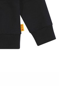 Schwarzer Sweatshirt mit gerippten Bündchen, versehen mit einem kleinen gelben Etikett mit rotem Logo. Baumwollmischung, weiche Textur, minimalistisches Design.