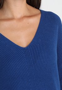 Maglione lavorato a maglia di un blu intenso con scollatura a V. Presenta coste testurizzate e maniche corte. Il tessuto sembra spesso e caldo.