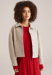 Beige uldblandet cropped jakke med foldet krave, frontlukning med knapper og to sidelommer. Bæres over en rød kjole.