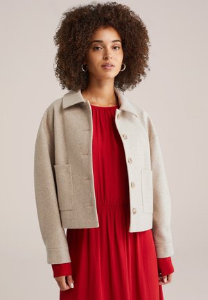 Beige Wollmischung Kürzer Jacke mit umgeschlagenem Kragen, vorderem Knopfverschluss und zwei seitlichen Taschen. Über einem roten Kleid getragen.