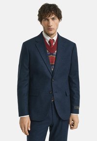 Marineblauer strukturierter Anzugjacke mit zwei Knöpfen, begleitet von einem rot gemusterten Pullover und einem weißen Hemd mit Kragen. Eine rote Krawatte rundet das Outfit ab.