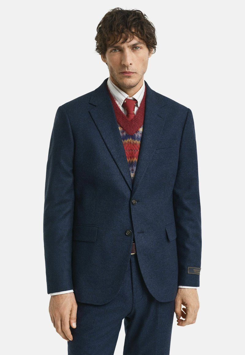 Marineblauer strukturierter Anzugjacke mit zwei Knöpfen, begleitet von einem rot gemusterten Pullover und einem weißen Hemd mit Kragen. Eine rote Krawatte rundet das Outfit ab.