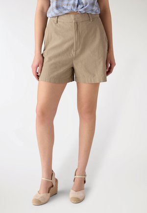 Donna con pantaloncini beige a vita alta, camicia a quadri blu annodata e sandali con zeppa beige, in piedi davanti a uno sfondo semplice.