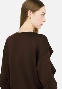 Brązowy oversize'owy sweter o prążkowanej fakturze, z szerokimi rękawami. Złote geometryczne kolczyki uzupełniają całość.