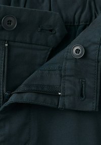 Gros plan sur un tissu noir avec une fermeture éclair partiellement dézippée, deux boutons noirs et leurs boutonnières correspondantes sur des couches de vêtements pliés.