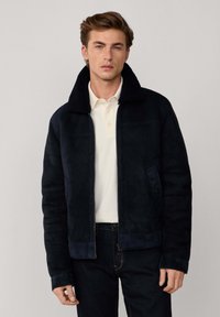 Marineblauwe suède jas met een zwarte schapenvachten kraag, ritssluiting, zijnaden en twee voorkanten zakken, gedragen over een witte polo.