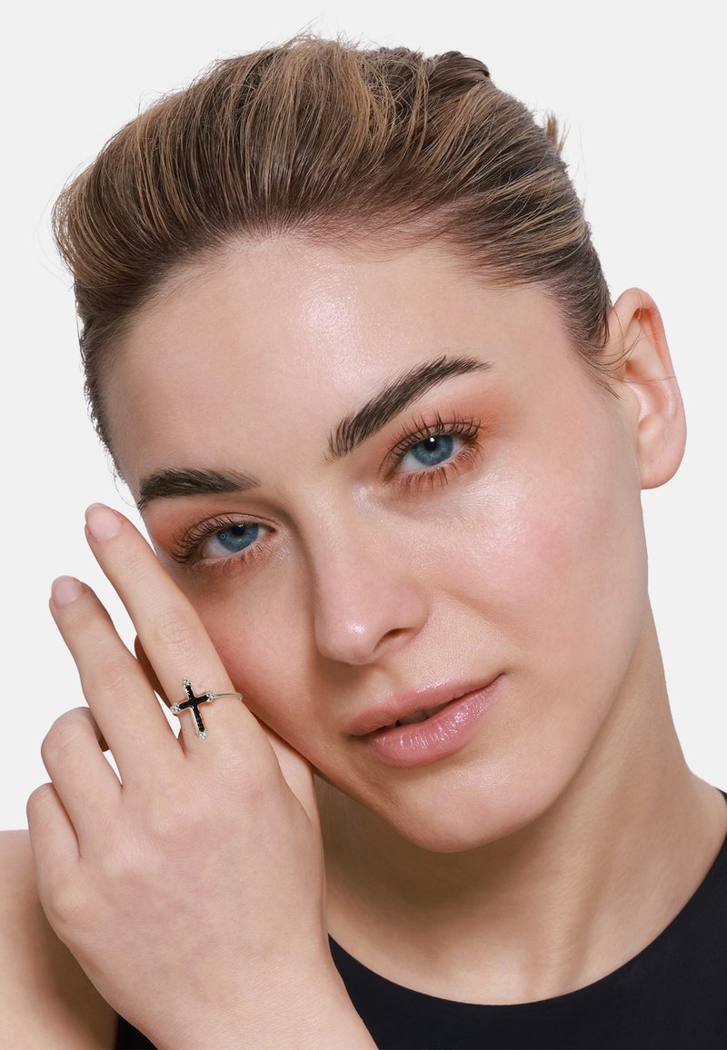 Srebrny pierścionek z czarnym wzorem krzyża. Modelka prezentuje pierścionek na palcu, ukazując gładką teksturę na tle neutralnego koloru skóry.