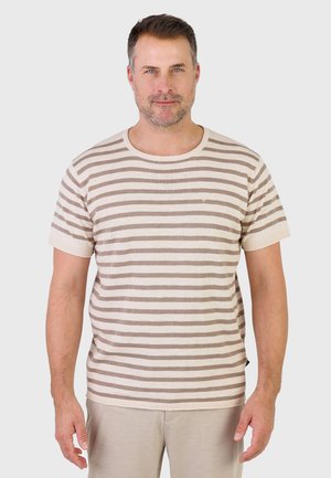 Uomo con capelli castani corti e barba chiara che indossa una camicia a maniche corte beige e bianca a strisce e pantaloni beige, in piedi davanti a uno sfondo semplice.