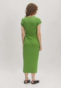 Grünes, knöchellanges Kleid mit tailliertem Oberteil, kurzen Fledermausärmeln und einem gedrehten Detail im Rücken. Glattes, leichtes Material. Schwarze Sandalen sichtbar.