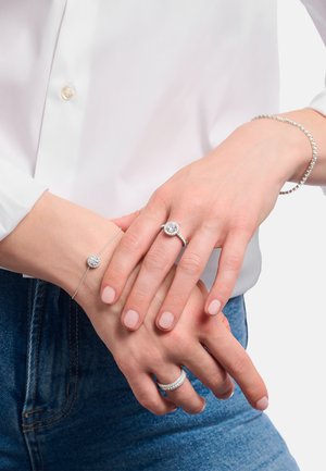 Verschränkte Hände zeigen silbernen Schmuck, darunter einen Diamantring, ein Armband mit rundem Anhänger und ein Tennisarmband, getragen von einer Person in weißem Oberteil und blauen Jeans.