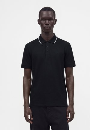 Homme portant un polo noir avec un liseré blanc au col, se tenant devant un fond blanc uni.
