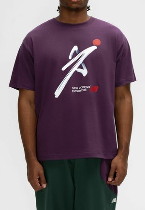 Mann trägt ein lila New Balance Basketball-T-Shirt mit einer weißen abstrakten Figur und rotem Punkt vorne, kombiniert mit grünen Shorts.