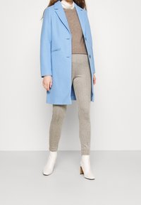 Hellblauer Wollmantel mit Reverskragen, kombiniert mit einem braunen Pullover und Hahnentritt-Musterhosen; weiße Ankle-Boots runden das Outfit ab.